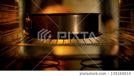 Brown cake bakes in a round pan inside an oven at home during the evening hours Brown cake bakes in a round pan inside an oven at home during the evening hours 138169814