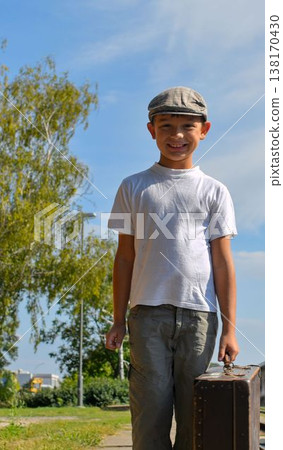 A cute boy at the train station. A little smiling boy holds an old suitcase. The concept of travel, vacation, and carefree childhood. Retro and vintage vibe. A cute boy at the train station. A little smiling boy holds an old suitcase. The concept of travel, vacation, and carefree childhood. Retro and vintage vibe. 138170430