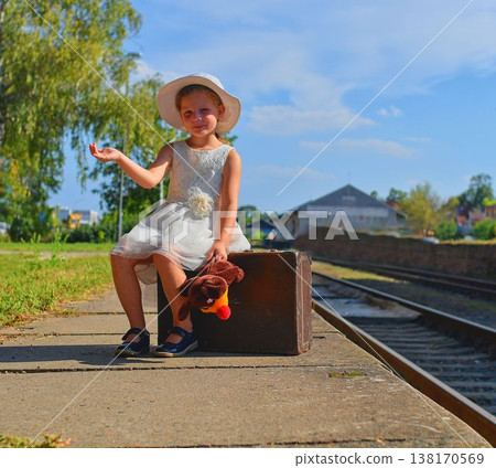 A lovely girl at the train station. A charming girl at the train station. A little girl holds a cuddly toy and sits on an old suitcase. The concept of travel, summer vacation, and a carefree A lovely girl at the train station. A charming girl at the train station. A little girl holds a cuddly toy and sits on an old suitcase. The concept of travel, summer vacation, and a carefree 138170569
