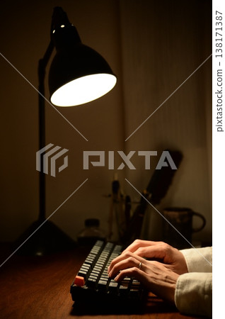 A man's hands typing on a keyboard while working remotely at night. A man's hands typing on a keyboard while working remotely at night. 138171387