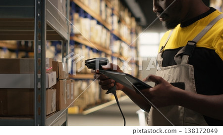African american man using scanner to register barcodes tags in inventory, solving logistics operations in industrial warehouse. Worker checking shipping labels near storage racks. Camera B. 138172049