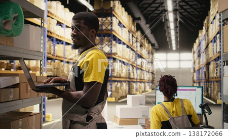 Black male staff verifying inventory pallets in e-commerce logistics center. Accurate order processing on laptop and packaging for quick shipping in large scale warehouse setting. Camera A. 138172066