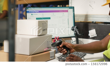 African American male employee scanning packages in a large warehouse, registering airway bills shipment details within industrial depot records. Using scanner for order fulfillment. Camera B. 138172096