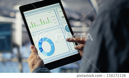 Close up of data center employee checking infrastructure diagnostics graphs on tablet screen. IT specialist on industrial platform in server room examining data charts during system audit, camera B Close up of data center employee checking infrastructure diagnostics graphs on tablet screen. IT specialist on industrial platform in server room examining data charts during system audit, camera B 138172136