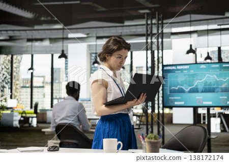 White woman accountant stands in office holding laptop, preparing budget report before board meeting. Focused female team leader uses digital device for business project planning. White woman accountant stands in office holding laptop, preparing budget report before board meeting. Focused female team leader uses digital device for business project planning. 138172174