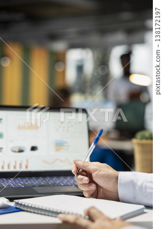 Close up of caucasian male hands taking notes with pen and analyzing financial graphs on digital device. Businessman reviews information on laptop and writes strategy plans on notebook. Close up of caucasian male hands taking notes with pen and analyzing financial graphs on digital device. Businessman reviews information on laptop and writes strategy plans on notebook. 138172197