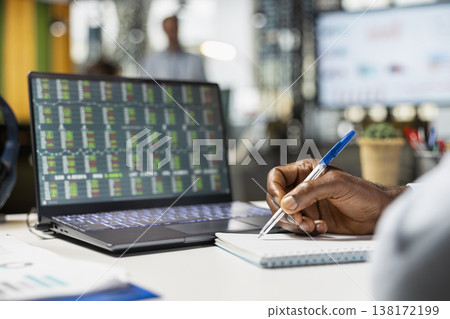 Close up of African American professional reviewing forex data and taking notes on paper, preparing strategy report. Black male individual writing information while analyzing trading charts on laptop. Close up of African American professional reviewing forex data and taking notes on paper, preparing strategy report. Black male individual writing information while analyzing trading charts on laptop. 138172199