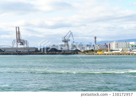 Industrial harbor with large cargo cranes, warehouses, and coal piles under a cloudy sky. Panoramic view of a commercial port facility across blue water with mountain range in the distance. Industrial harbor with large cargo cranes, warehouses, and coal piles under a cloudy sky. Panoramic view of a commercial port facility across blue water with mountain range in the distance. 138172957