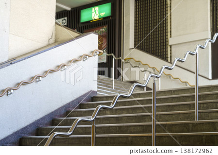 Modern ergonomic wavy handrails on public stairs in Japan. Unique zigzag safety railing design for better grip and accessibility in an urban station. Emergency exit sign visible in the background. 138172962
