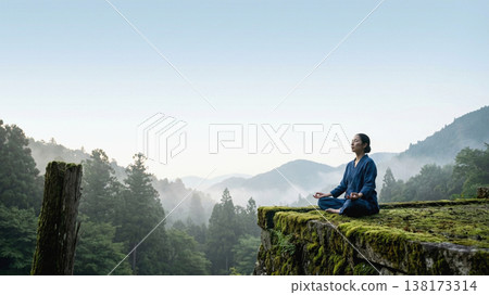A woman meditating amidst the tranquility of the deep mountains; a landscape of moss-covered stone walls and morning mist. 138173314