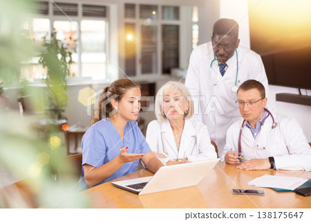 Doctor with colleagues sitting at table, reading new of law on laptop and discussing information 138175647