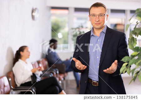 Dispassionate secretary man stands at reception with folder and list of guests and expects visitors Dispassionate secretary man stands at reception with folder and list of guests and expects visitors 138175845