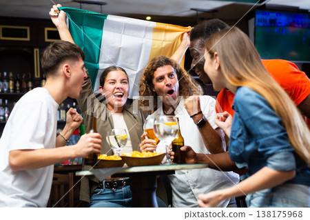 Happy international football supporters holding up the flag of Ireland and drinking beer in the pub Happy international football supporters holding up the flag of Ireland and drinking beer in the pub 138175968