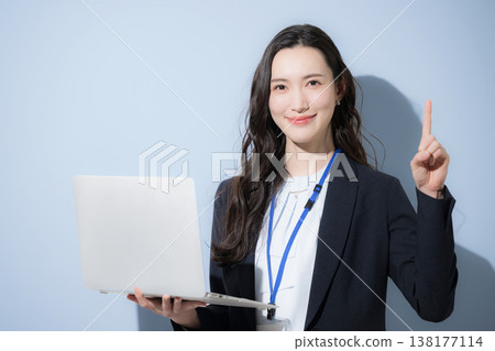 一位身著職業套裝、佩戴名牌、手持筆記型電腦的時尚女性出現在淺藍色背景前,她正對著鏡頭,並用手指著什麼(僅上半身)。 一位身著職業套裝、佩戴名牌、手持筆記型電腦的時尚女性出現在淺藍色背景前,她正對著鏡頭,並用手指著什麼(僅上半身)。 138177114