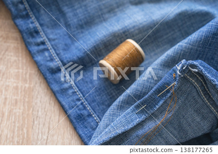 Denim fabric with needle, wooden thread spool and pins arranged for mending and tailoring a blue jean garment, close-up workshop scene. Repairing denim clothing with needle and thread Denim fabric with needle, wooden thread spool and pins arranged for mending and tailoring a blue jean garment, close-up workshop scene. Repairing denim clothing with needle and thread 138177265