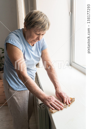 Senior woman performing domestic chores, carefully wiping dust from a window surface during daily routine Senior woman performing domestic chores, carefully wiping dust from a window surface during daily routine 138177293