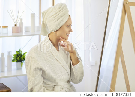 Smiling woman in bathrobe with towel on head enjoying fresh skin and looking at mirror after shower. Smiling woman in bathrobe with towel on head enjoying fresh skin and looking at mirror after shower. 138177331