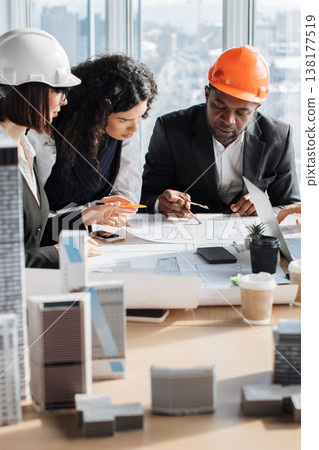 A diverse team of architects and engineers collaborates around a table, reviewing construction plans and building models with focused attention A diverse team of architects and engineers collaborates around a table, reviewing construction plans and building models with focused attention 138177519