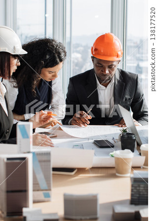 A diverse team of architects wearing hard hats collaborates around a table, reviewing construction plans and building models A diverse team of architects wearing hard hats collaborates around a table, reviewing construction plans and building models 138177520