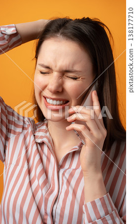Young woman talks on phone with a frustrated expression against a bright orange background 138177710