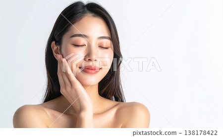 一位年輕的日本女性面帶微笑,輕撫臉頰檢查肌膚水分,象徵護膚和美麗的肌膚。 一位年輕的日本女性面帶微笑,輕撫臉頰檢查肌膚水分,象徵護膚和美麗的肌膚。 138178422