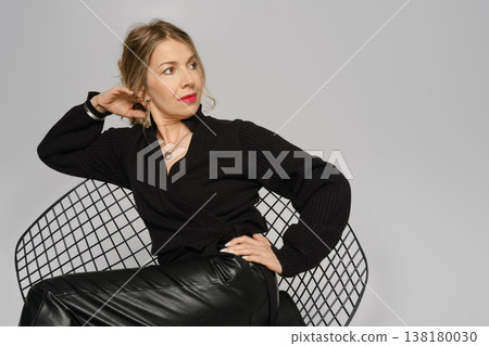 Woman posing on chair in studio, focused on direction, wearing black top and leather pants 138180030
