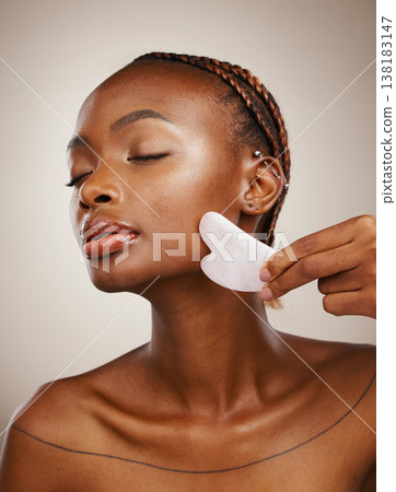 Woman, beauty and gua sha for face massage, aesthetic skincare and anti aging dermatology in studio on grey background. African model, crystal stone tools or lymphatic drainage with facial cosmetics Woman, beauty and gua sha for face massage, aesthetic skincare and anti aging dermatology in studio on grey background. African model, crystal stone tools or lymphatic drainage with facial cosmetics 138183147