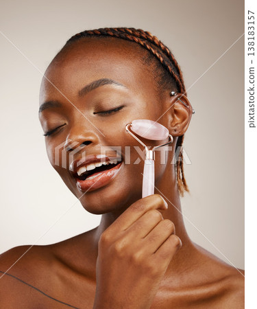 Woman, face roller and massage in studio for skincare, rose quartz cosmetics and aesthetic dermatology on brown background. African model, crystal stone tools and lymphatic drainage for facial beauty 138183157