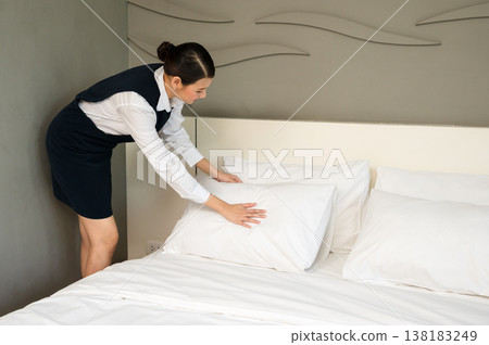 Hotel housekeeper in uniform neatly arranging white pillows on freshly made bed in clean modern guest room Hotel housekeeper in uniform neatly arranging white pillows on freshly made bed in clean modern guest room 138183249