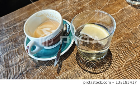 Coffee cup featuring latte art on a blue saucer next to a glass of water with lemon slice on a wooden table in a cafe environment Coffee cup featuring latte art on a blue saucer next to a glass of water with lemon slice on a wooden table in a cafe environment 138183417