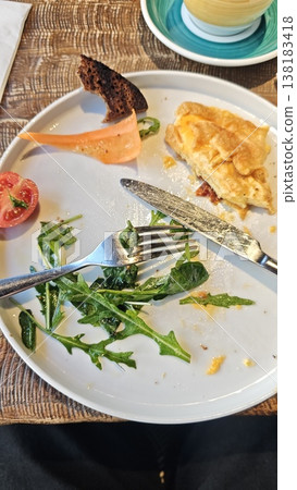 Plate with remnants of a meal featuring omelette, salad, tomato, and bread, accompanied by a fork and knife on a textured table surface 138183418