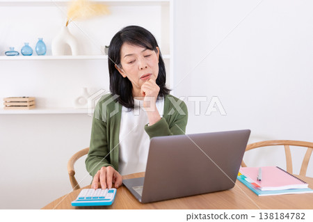 A housewife in her 50s struggling with a computer and calculator. A housewife in her 50s struggling with a computer and calculator. 138184782
