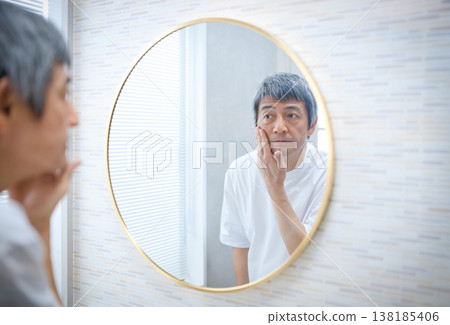 A man in his 60s touches his face with his hands while looking in a mirror. 138185406