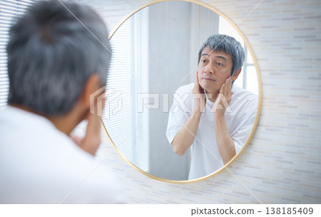 一位六十多歲的男子對著鏡子，用手撫摸自己的臉。 138185409