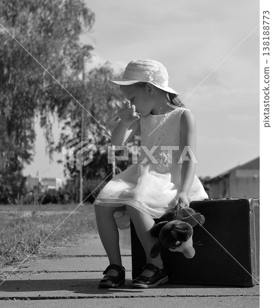 Young girl in a white dress and hat sits on a suitcase with a toy, looking sad Young girl in a white dress and hat sits on a suitcase with a toy, looking sad 138188773