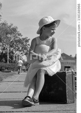 Young girl in a hat sits on an old suitcase holding a pillow, looking away Young girl in a hat sits on an old suitcase holding a pillow, looking away 138188966