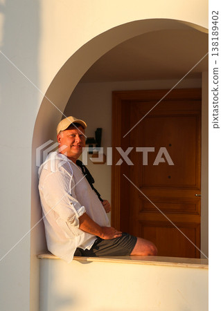 Adult man in a white shirt and cap sitting on a wide windowsill, enjoying the beautiful sunset, reflecting on life and tranquility in the golden hour 138189402