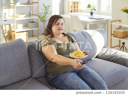 Fat overweight woman sitting on sofa at home watching tv and eating tasty french fries. Fat overweight woman sitting on sofa at home watching tv and eating tasty french fries. 138191935