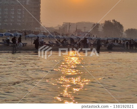 Golden Sunset Over Crowded Beach With Sun Reflection on Water and City Silhouette Golden Sunset Over Crowded Beach With Sun Reflection on Water and City Silhouette 138192021