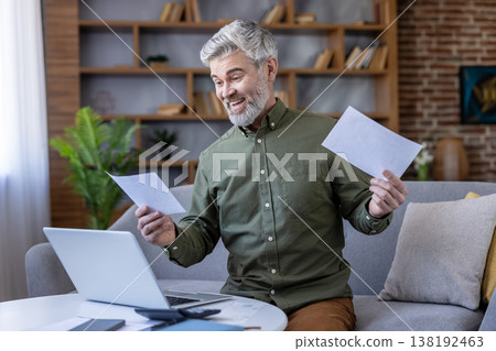 Senior man smiling excitedly while reviewing paperwork and using a laptop at a home office setup, celebrating financial success and managing bills online 138192463