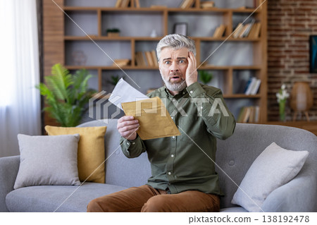 Mature man sitting on a sofa in his living room, opening a letter and reacting with shock and worry upon reading bad news, symbolizing financial difficulty and unexpected problems Mature man sitting on a sofa in his living room, opening a letter and reacting with shock and worry upon reading bad news, symbolizing financial difficulty and unexpected problems 138192478