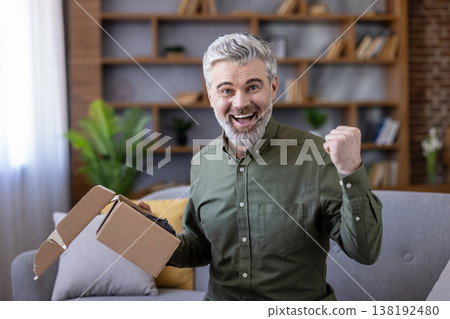 Excited senior man is holding an open cardboard box, showing joy and satisfaction after receiving a parcel from an online order while sitting on a couch at home Excited senior man is holding an open cardboard box, showing joy and satisfaction after receiving a parcel from an online order while sitting on a couch at home 138192480