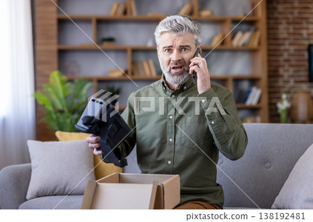 Mature man receiving a damaged virtual reality headset from an online delivery, looking frustrated while talking on a mobile phone to customer service in his living room Mature man receiving a damaged virtual reality headset from an online delivery, looking frustrated while talking on a mobile phone to customer service in his living room 138192481