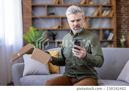 Mature man holding an open cardboard box and smartphone, looking disappointed and stressed after receiving an incorrect or damaged online order at home Mature man holding an open cardboard box and smartphone, looking disappointed and stressed after receiving an incorrect or damaged online order at home 138192482