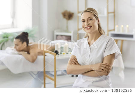 Portrait of smiling female massage therapist in white uniform posing in spa room with client behind. 138192556