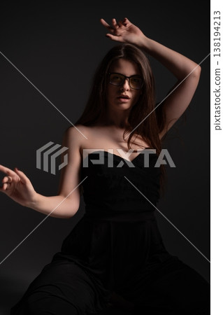 Woman wearing a sleek black strapless jumpsuit and modern clear glasses strikes a fashionable pose with her arms, exuding confidence and elegance in a studio setting Woman wearing a sleek black strapless jumpsuit and modern clear glasses strikes a fashionable pose with her arms, exuding confidence and elegance in a studio setting 138194213