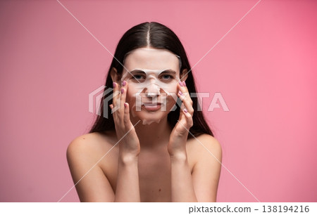 Woman applying a hydrating sheet mask on her face for a refreshing beauty treatment and self-care routine, promoting healthy skin and wellness against a pink background 138194216