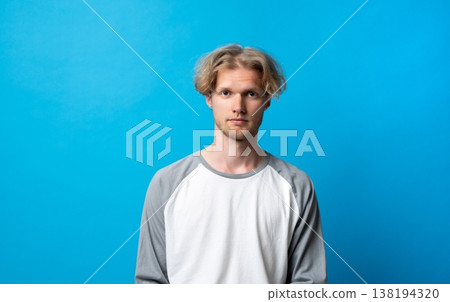 Young blond man staring seriously at camera, showing calm expression and confident attitude, standing in front of a vibrant blue background, embracing youth and casual style Young blond man staring seriously at camera, showing calm expression and confident attitude, standing in front of a vibrant blue background, embracing youth and casual style 138194320