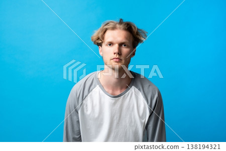Young man with blond hair and a neutral expression standing directly facing the camera, wearing a casual long-sleeve t-shirt against a striking blue studio backdrop Young man with blond hair and a neutral expression standing directly facing the camera, wearing a casual long-sleeve t-shirt against a striking blue studio backdrop 138194321