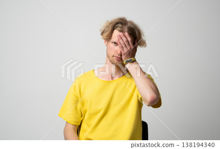 Young man wearing a yellow t-shirt partially covering his face with his hand, conveying concepts of shame, hiding, privacy, emotion, secret, and disappointment 138194340